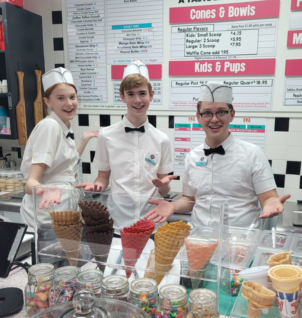 Three workers at an ice cream shop displaying colorful cones, with a menu in the background