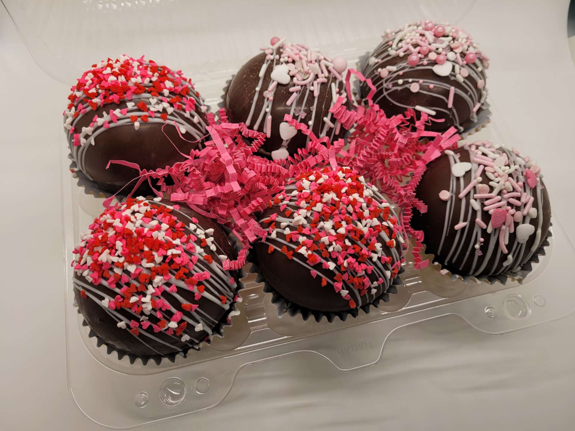 Six hot chocolate bombs, also known as hot cocoa bombs, decorated with red and white sprinkles and pink and white drizzle